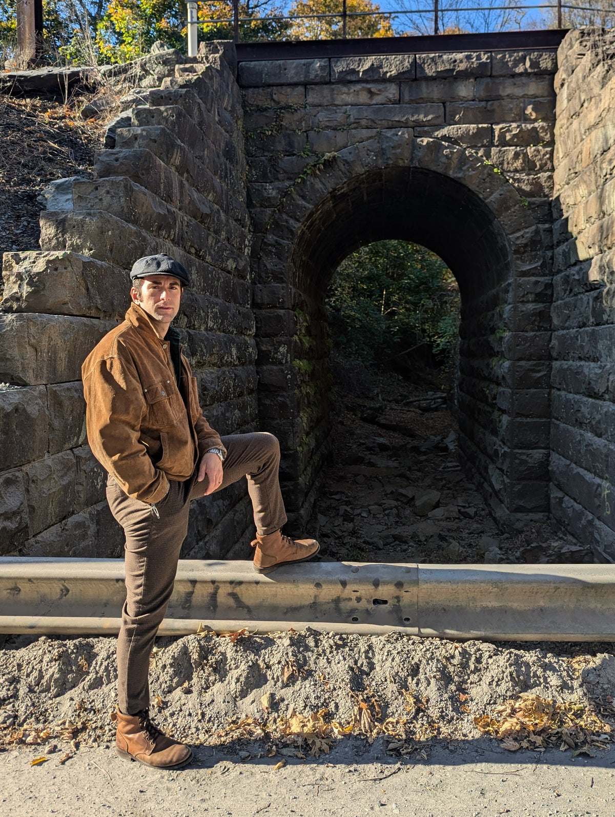 A man wearing a brown jacket, brown pants, and a gray flat cap stands on a concrete barrier in front of a large stone archway and tunnel structure outdoors under a clear blue sky.